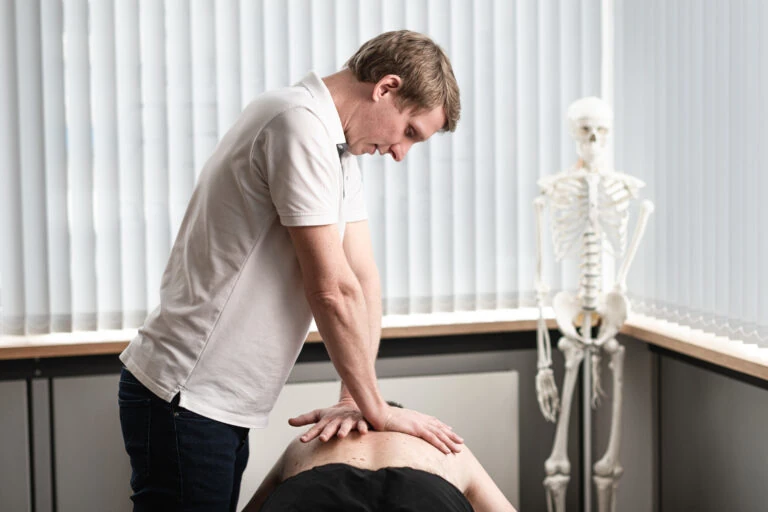 Physiotherapeut behandelt Patientenrücken in moderner Praxis, Skelettmodell im Hintergrund.