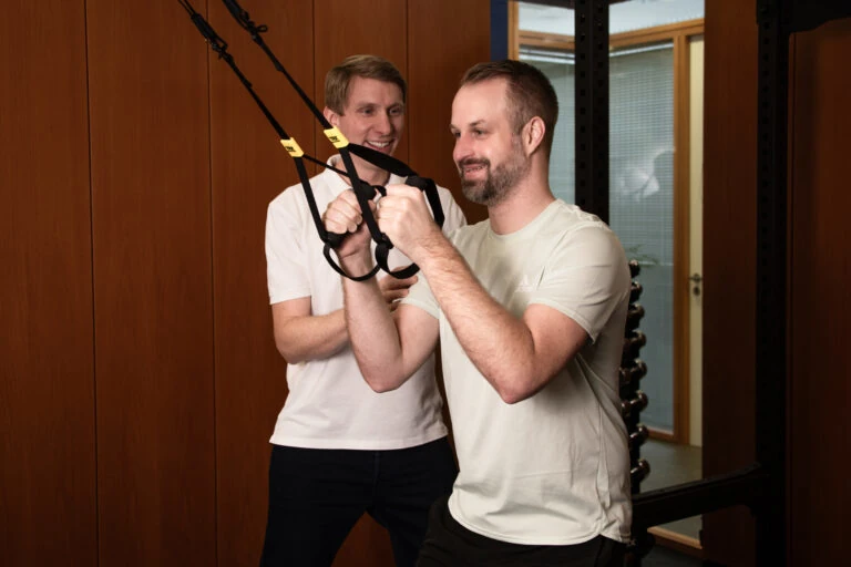 Zwei Männer trainieren gemeinsam mit einem TRX-Band in einem Fitnessraum.